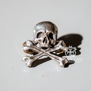 Könnte beinhalten: Eine silberne Totenkopf- und Knochen-Anstecknadel mit goldener Akzent. Die Anstecknadel ist ein Schmuckstück mit dem Text "IRON CLAN JEWELRY" auf der Unterseite.