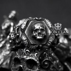 May include: A silver ring with a skull design. The skull is in the center of the ring and is surrounded by intricate details. The ring is made by Iron Clan Jewelry.
