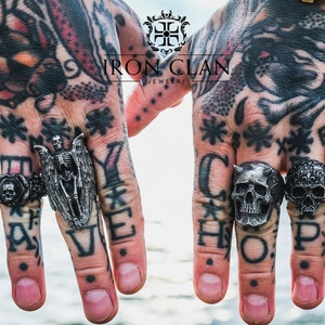 May include: Two hands wearing silver rings with skull and skeleton designs. The rings are from Iron Clan Jewelry and the hands are tattooed. The rings spell out "Stay" and "Hope".