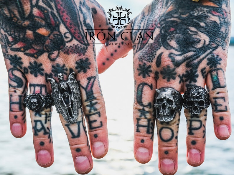May include: Two hands wearing silver rings with skull and skeleton designs. The rings are from Iron Clan Jewelry and the hands are tattooed. The rings spell out "Stay" and "Hope".