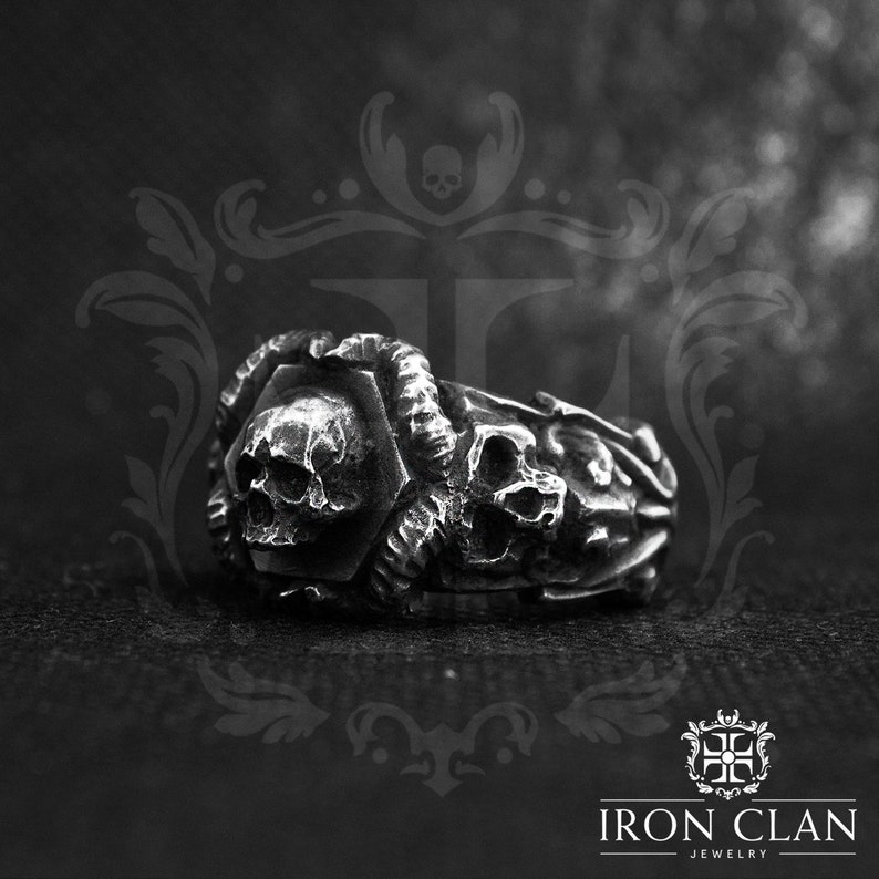 May include: A silver skull ring with intricate details. The ring features a large skull in the center, surrounded by smaller skulls and other decorative elements. The ring is made of high-quality metal and has a polished finish.