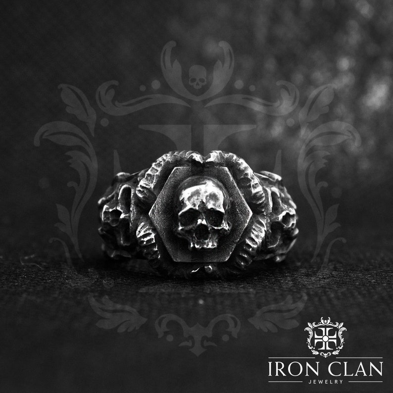 May include: A silver ring with a skull design. The skull is surrounded by a hexagonal frame and is flanked by two snake-like figures. The ring is made by Iron Clan Jewelry.