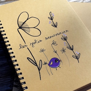 Puede incluir: Cuaderno con espiral y cubierta de papel kraft. La portada presenta un diseño dibujado a mano de una flor grande, un pájaro morado y elementos florales. Las palabras "les jolis souvenirs" están escritas en cursiva. El cuaderno está abierto a páginas en blanco.