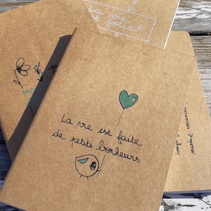 Puede incluir: Varios cuadernos de papel kraft con frases e ilustraciones manuscritas en francés. Un cuaderno presenta un globo con forma de corazón verde y la frase "La vie est faite de petits bonheurs".