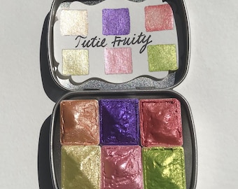 May include: A metal tin containing six watercolour pans. The pans are arranged in two rows of three. The colours are: gold, purple, pink, green, gold, pink, and green. The tin has the words "Tutie Fruity" written on it.