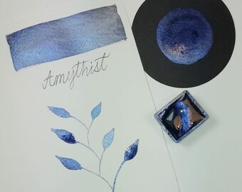 May include: A watercolor paint pan with a blue and silver shimmery paint. The paint is labeled "Amethyst" and a sample of the paint is shown on a white card with a blue and silver shimmery finish. A small sample of the paint is also shown on a black card.