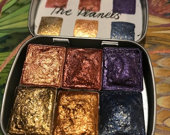 May include: A metal tin with six watercolor paint pans. The pans are filled with metallic paints in shades of gold, copper, and purple. The lid of the tin has the words "The Planets" written in black ink.