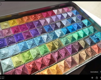 May include: A metal tin filled with small squares of watercolor paint. The paint is arranged in rows and columns, and each square has a different color. The colors are vibrant and range from deep blues and greens to bright pinks and oranges.