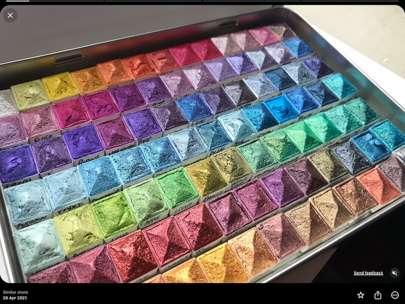 May include: A metal tin filled with small squares of watercolor paint. The paint is arranged in rows and columns, and each square has a different color. The colors are vibrant and range from deep blues and greens to bright pinks and oranges.