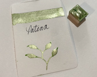 Peut inclure: Une carte d'échantillon de peinture aquarelle avec le mot "Patena" en écriture noire et une branche de feuilles vert métallisé. Un petit godet de peinture carré de la même couleur est à proximité. La carte a une bande verte métallique en haut.