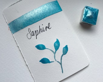 May include: A small, square, teal blue watercolor paint pan with the word "Sapphire" written in black ink on a white card. The card also has a teal blue watercolor stripe and a small teal blue watercolor leaf design.