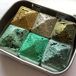 May include: A set of six metallic watercolor paints in a small, rectangular tin. The colors include gold, silver, and various shades of green. Each paint is a square shape and sits on a white base. The tin is open, revealing the paints.