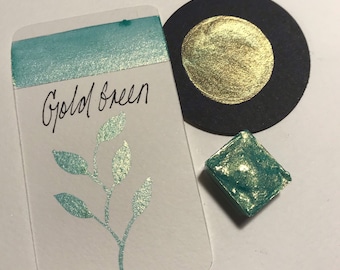 Pode incluir: Uma pequena bandeja quadrada de tinta aquarela verde-azulada com um brilho dourado. A tinta é etiquetada como "Gold Green" em um cartão branco com um desenho de folha verde.