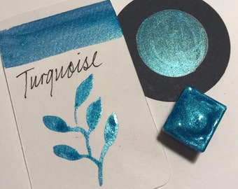 May include: A turquoise watercolor paint pan with a metallic sheen. The paint is in a small square pan and is labeled "Turquoise".