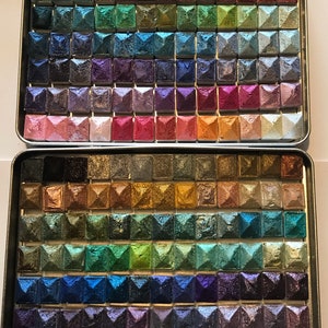 May include: A metal palette containing 100 small squares filled with different coloured pigments. The pigments are arranged in rows and columns, and each square has a raised, pyramid-shaped top. The palette is open and the pigments are visible.