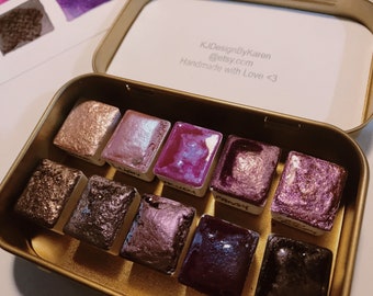 May include: A set of 10 watercolor paints in a gold metal tin. The paints are in small square pans and have a variety of shimmery colors, including gold, pink, purple, and brown. The tin has the text "KJDesignByKaren @etsy.com Handmade with Love <3" on the lid.