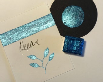 May include: A collection of shimmering blue watercolor samples. Includes a square, a circle, and a rectangular swatch with the word "Ocean" and a leaf design. The samples are on a white surface.
