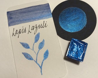 May include: A watercolor paint sample card with the text "lapis lazuli" written in cursive. The card shows a blue watercolor wash and a blue leaf design. A small blue paint pan and a round blue paint sample are also shown.