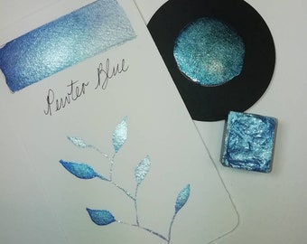 May include: A white card with a blue watercolor swatch and the handwritten text "Pewter Blue". A black circle with a blue shimmer and a small square of blue watercolor are also shown. A silver and blue leaf design is on the card.