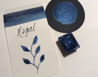 May include: A collection of watercolor paints in a deep blue hue. The image includes a swatch on a white card with the word "Regal" and a botanical illustration, a circular paint sample, and a square paint pan.