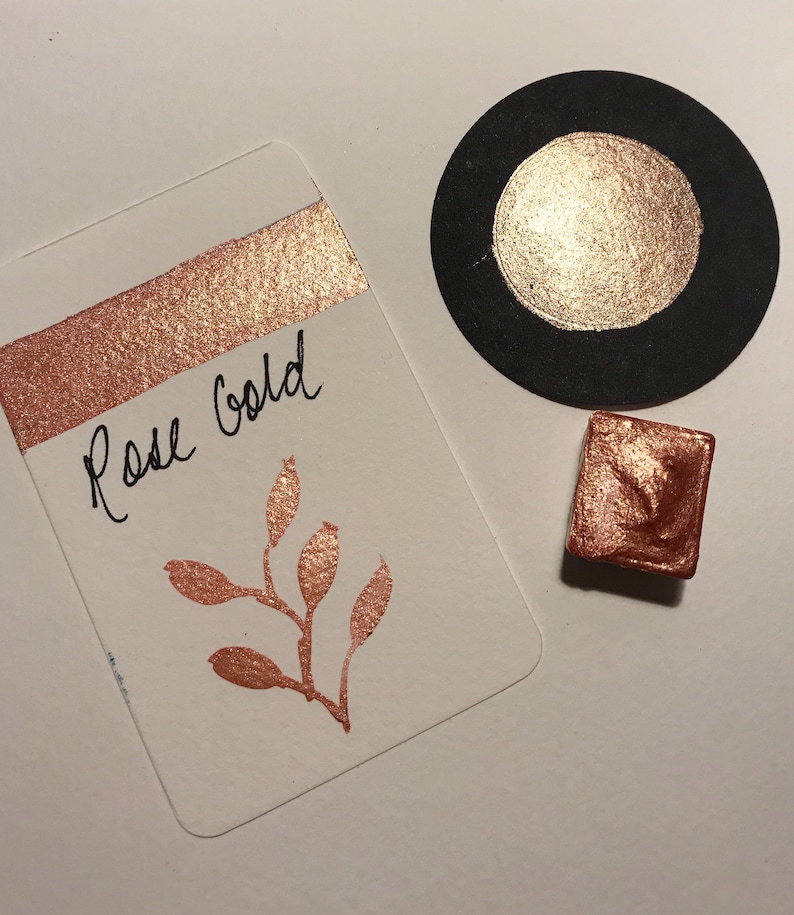 Puede incluir: Una peque&ntilde;a bandeja cuadrada de pintura de acuarela de oro rosa. La bandeja est&aacute; sobre una superficie blanca junto a una tarjeta con las palabras "Rose Gold" escritas en ella. La tarjeta tambi&eacute;n tiene un peque&ntilde;o dise&ntilde;o de oro rosa de una rama con hojas y bayas.