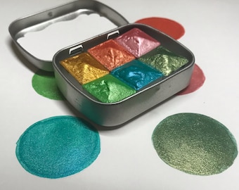 May include: A metal tin containing six pans of metallic watercolour paint. The pans are arranged in a 2x3 grid. The colours are gold, orange, pink, green, teal, and a darker green. The tin is open and the paint is visible. The paint is arranged in a circle on a white surface.