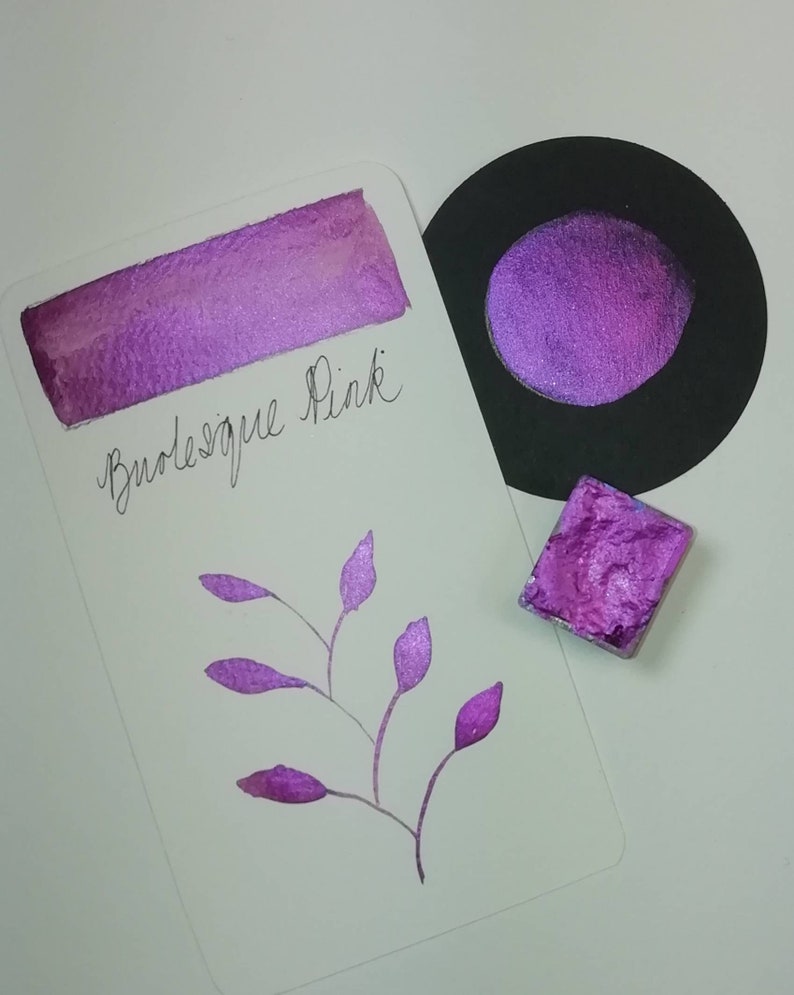 May include: A purple watercolor paint pan with a shimmery finish. The paint is labeled "Burlesque Pink".