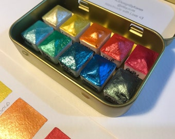 May include: A set of 10 metallic watercolor paints in a gold tin. The paints are arranged in a rainbow order, with the colors being: silver, gold, teal, green, blue, turquoise, teal, red, and purple.