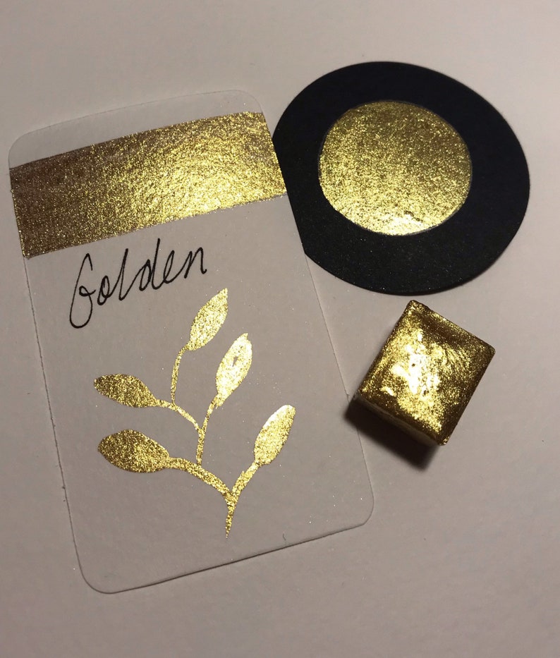 May include: A gold watercolor paint pan with a gold leaf finish. The pan is labeled "Golden" and is surrounded by a gold leaf swatch and a gold leaf circle on black paper.