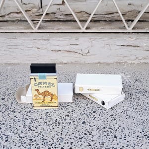 May include: A pack of Camel Filter cigarettes with a camel illustration and the text "TURKISH & AMERICAN BLEND" on the front. The pack is sitting on a white surface.