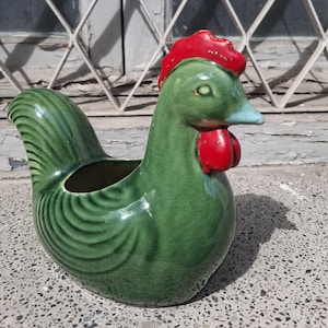 Pode incluir: Um vaso de cerâmica em forma de galo com acabamento verde brilhante e textura de esmalte craquelado. O galo apresenta uma crista e barbelas vermelho-vivas, um bico azul-claro e uma abertura oca nas costas para colocar pequenos objetos.
