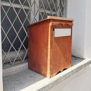 Puede incluir: Un buzón de madera con acabado natural. Tiene forma rectangular, con una parte superior decorativa y una ranura metálica para el correo. Está sobre una repisa de piedra, con una ventana con una reja metálica en forma de diamante en el fondo.