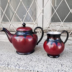 May include: A vintage tea set featuring a maroon teapot with a black handle and lid, and a matching creamer. The set has a black and maroon design and is placed on a speckled surface.