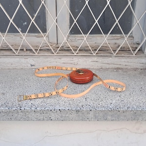 Puede incluir: Una cinta métrica naranja y marrón vintage con una caja circular. La cinta está extendida, mostrando marcas en centímetros. La cinta métrica está sobre una superficie gris, con una ventana y una rejilla metálica blanca en el fondo.