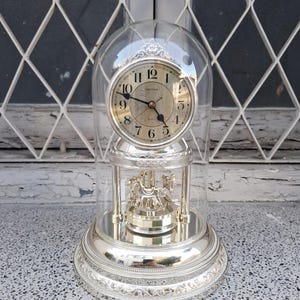May include: A gold-tone Rhythm mantel clock encased in a clear glass dome. The clock features a round face with black Arabic numerals and hands, sitting above a rotating carousel base with metallic horse figures on a circular mirrored platform.