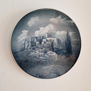 Puede incluir: Un plato decorativo redondo con una pintura en escala de grises del Partenón sobre una colina, con nubes en el cielo. El plato tiene un borde azul oscuro y está montado en una pared blanca.
