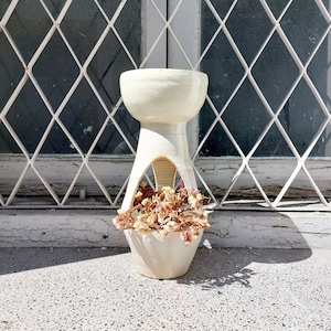 May include: White ceramic planter with a unique design featuring a wide opening at the top and a narrow opening at the bottom. The planter is filled with dried flowers.