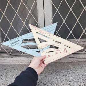Könnte beinhalten: Zwei Vintage-Zeichenwerkzeuge, ein hellblaues und ein cremefarbenes Dreieckslineal, werden vor einem Fenster gehalten. Das hellblaue Lineal trägt den Text "DAVANTI DEVANT". Das cremefarbene Lineal trägt den Text "ARMEL".
