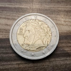 Può includere: Una moneta da due euro con un ritratto di profilo di Dante Alighieri che indossa una corona d'alloro. La moneta ha un centro color oro e un anello esterno color argento con stelle. Il testo IR, 2002, R e MCC è visibile sulla faccia.