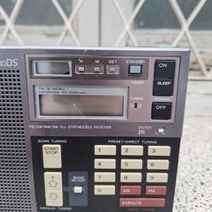 May include: A close-up of a vintage gray radio with a digital display and numerous buttons. The radio features buttons for power, tuning, and preset channels. The text on the radio includes "FM 76-108 MHz" and "FM/LW/MW/SW PLL SYNTHESIZED RECEIVER."