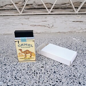 Puede incluir: Un paquete de cigarrillos vintage Camel Filters con una ilustraci&oacute;n de camello y el texto "Turkish & American Blend Cigarettes". El paquete est&aacute; cerrado y se encuentra sobre una superficie blanca.