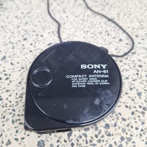 May include: Black Sony AN-61 compact antenna with attached hanger clip and reel-in carrying case. The antenna is round with a black cord attached. Text on the antenna reads "COMPACT ANTENNA FOR SHORT WAVE".