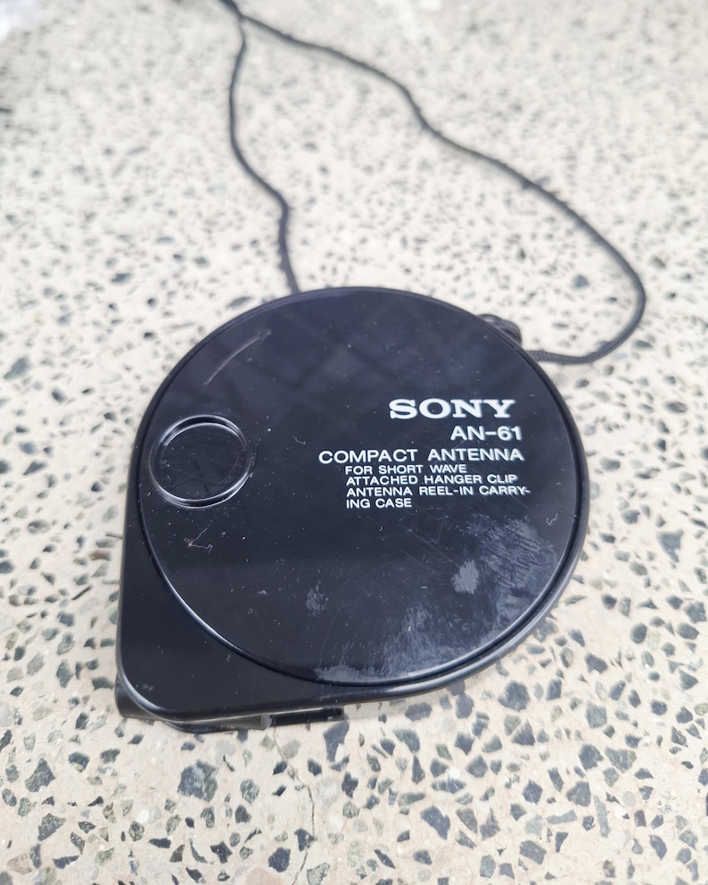 May include: Black Sony AN-61 compact antenna with attached hanger clip and reel-in carrying case. The antenna is round with a black cord attached. Text on the antenna reads "COMPACT ANTENNA FOR SHORT WAVE".