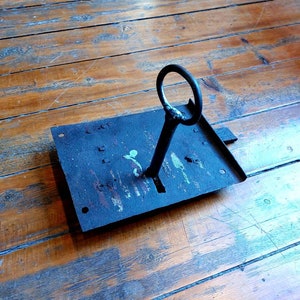May include: A vintage metal key with a circular handle, inserted into a black metal lock plate. The lock plate has a rectangular shape and is covered in rust and paint.