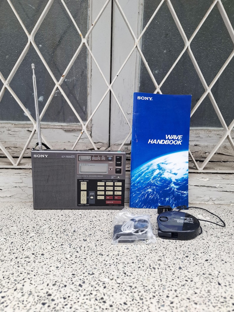 May include: A vintage Sony ICF-7600DS shortwave radio with a telescopic antenna, a blue "Wave Handbook", a black cassette player, and accessories. The radio is dark gray with a digital display and control buttons.