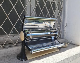 May include: A vintage electric heater featuring a curved chrome reflector, a central heating element protected by a horizontal wire cage, and a black and cream-colored base. The unit has a sleek, mid-century design with a power cord attached.