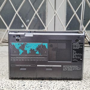 May include: A gray Sony ICF-7600DS radio receiver with a retractable antenna. The front panel features a world map graphic in teal, a frequency scale, and text including "TIME SET" and "RADIO BATTERY".