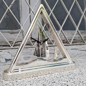 Può includere: Orologio al quarzo argentato in una cornice triangolare, con un design a barca a vela. Il quadrante ha lancette nere e piccoli punti neri per gli indicatori delle ore. La base è un rettangolo arrotondato. Il testo "RHYTHM QUARTZ JAPAN" è visibile.