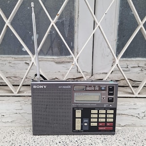 May include: A gray Sony ICF-7600DS portable radio with a retractable antenna. The radio has a digital display, a keypad, and various buttons for tuning and preset stations. The radio is set against a window with a white grid.
