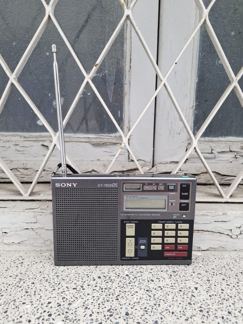 May include: A gray Sony ICF-7600DS portable radio with a retractable antenna. The radio has a digital display, a keypad, and various buttons for tuning and preset stations. The radio is set against a window with a white grid.